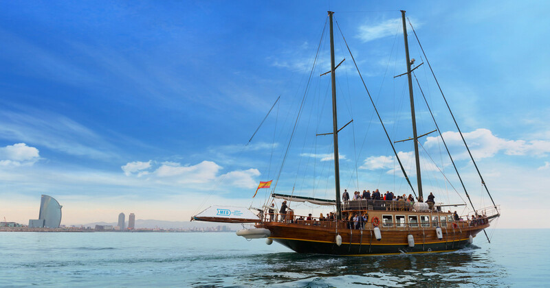 A traditional schooner sails along the calm waters of Barcelona's coastline, with the city's iconic skyline in the background, offering a unique perspective on the Catalan capital's scenic beauty and nautical charm.