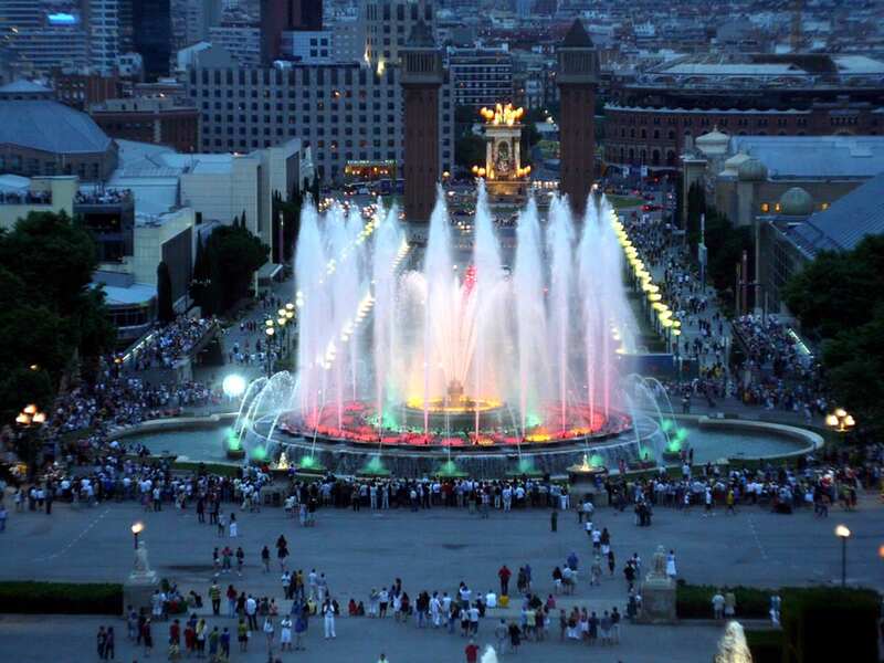 Twilight view of the Magic Fountain of Montjuïc in Barcelona, with colorful lights reflecting in the water as crowds gather to enjoy the spectacular display of choreographed water, light, and music.