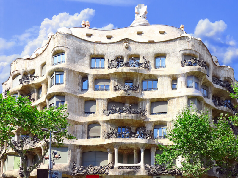The stunning façade of Casa Milà, also known as La Pedrera, one of Barcelona's most famous Modernist buildings by architect Antoni Gaudí, with visitors gathered outside to admire its unique, undulating stone exterior and wrought iron balconies against a vibrant blue sky.