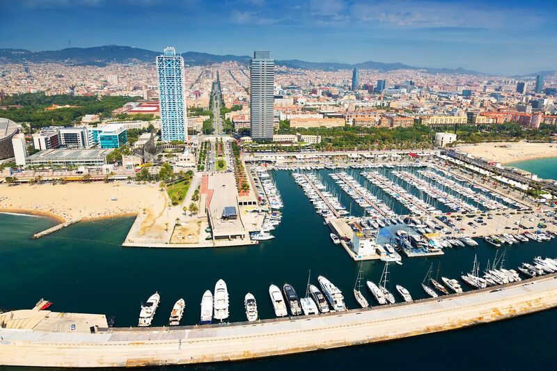 Aerial image of Port Olympic in Barcelona, showcasing its marina filled with yachts, the sandy beaches alongside, and the city skyline featuring iconic skyscrapers under a clear blue sky.