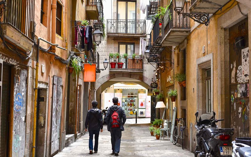 Two travelers exploring the narrow, cobblestone streets of the El Born District in Barcelona, passing by traditional balconies adorned with plants and historic facades with graffiti art, embodying the city's vibrant culture.