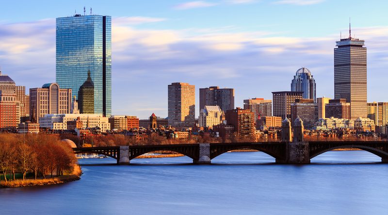 Sunset view of Boston's skyline with prominent skyscrapers casting reflections on the Charles River, the historic Longfellow Bridge in the foreground, and a backdrop of pastel-colored skies.