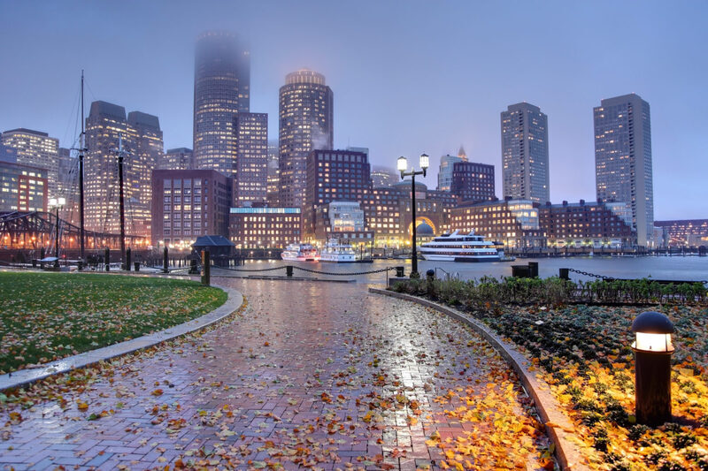 A serene dusk setting over Boston's waterfront, with a cobblestone path lined with fallen autumn leaves leading towards a backdrop of illuminated skyscrapers and moored boats, encapsulating the essential experiences to savor before visiting Boston.