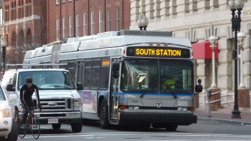 Photo of a Boston city bus with 'SOUTH STATION' displayed on its front, heading through a busy street. A cyclist is riding alongside, and cars are visible in the foreground. The urban environment suggests a typical day of transit in Boston, highlighting the city's commitment to accessible and varied transportation options for its residents and visitors.
