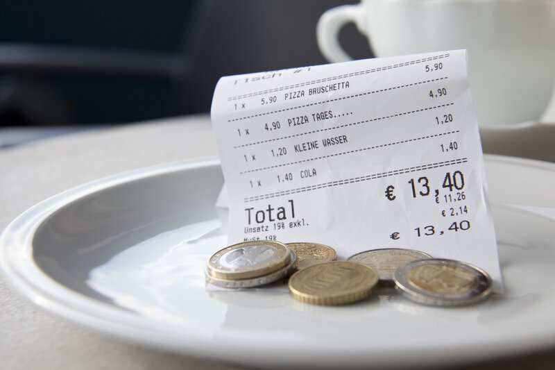 A close-up photo showing a restaurant bill and coins on a white plate, indicating a payment and tip left for service. The receipt includes items such as pizza and drinks, with a total of 13.40 euros, suggesting a dining experience. This image serves as a visual reference for the tipping practices important to know before visiting Boston.